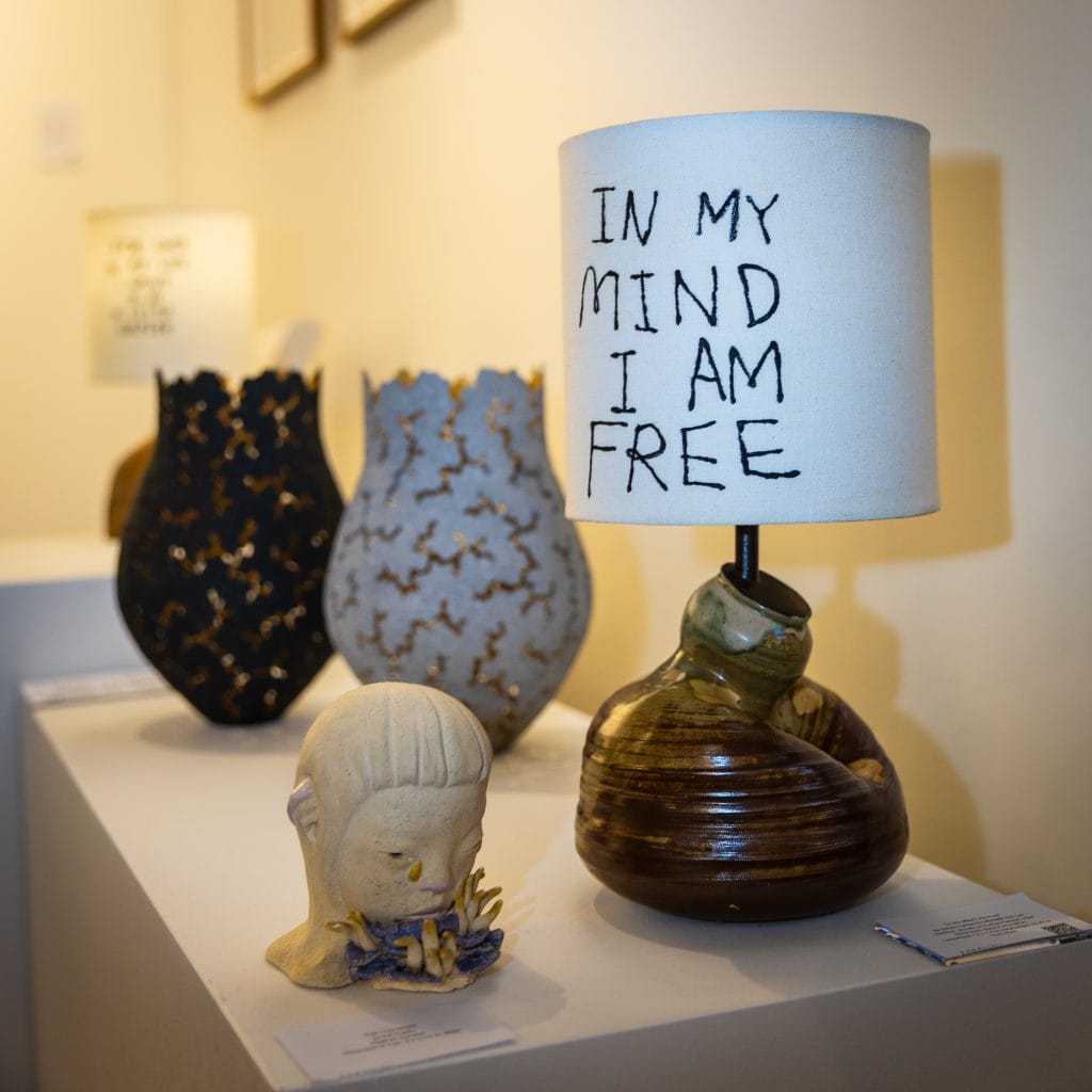 A display of ceramic artworks on a white pedestal in a gallery setting. In the foreground, a small sculpted head with closed eyes rests on a base decorated with organic shapes. Beside it is a brown, rounded ceramic lamp with a white shade that reads “IN MY MIND I AM FREE” in hand-drawn black lettering. In the background, two textured vases—one dark and one light—sit slightly out of focus, with another similar lamp further back. The lighting is warm and soft, highlighting the handmade textures. Featured at San Francisco Art Fair 2026.