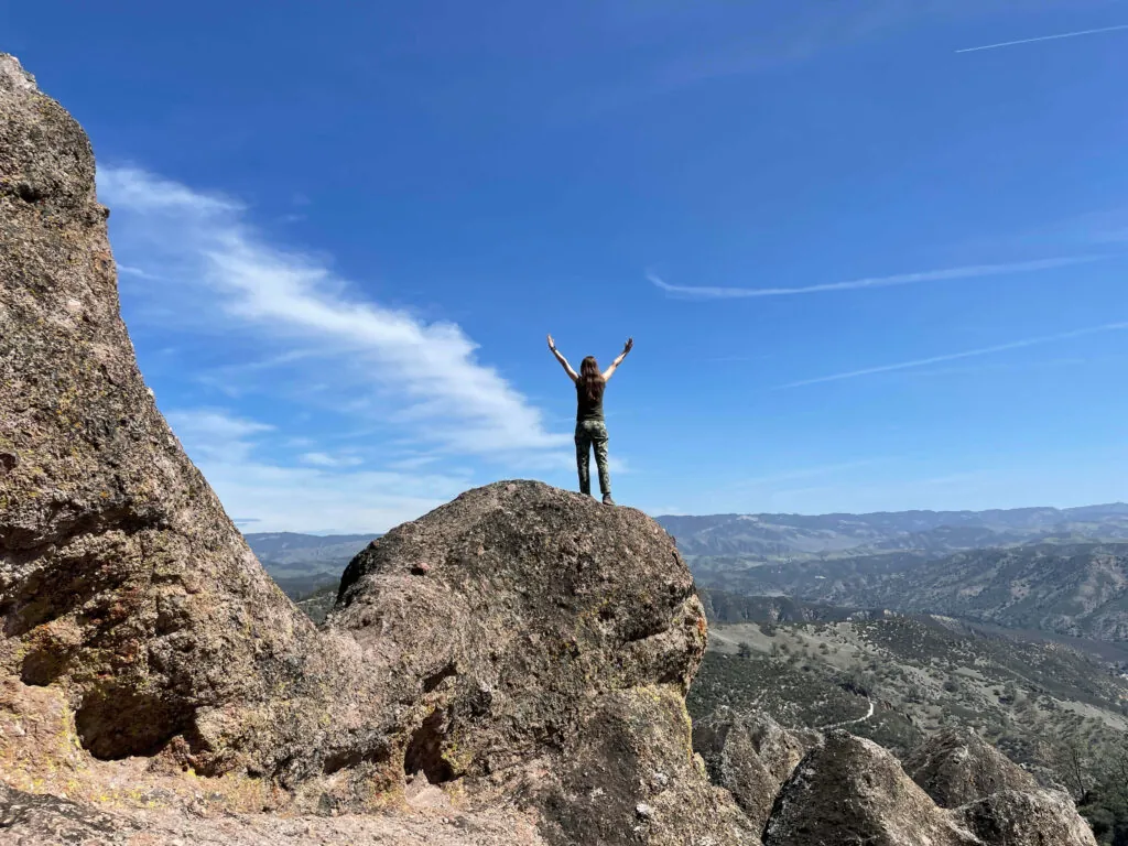 Person standing on a rocky cliff with arms raised, overlooking a vast mountain landscape