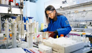 Scientist in a blue lab coat and safety glasses and black gloves working in a lab.