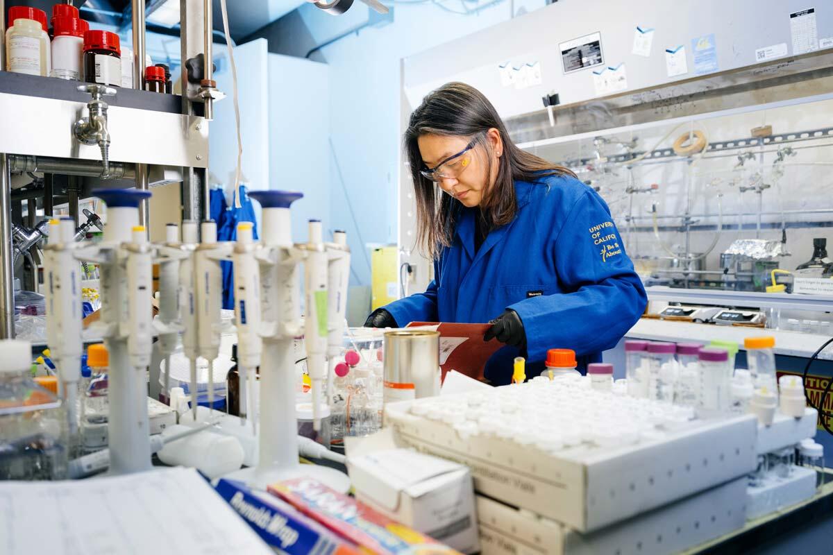 Scientist in a blue lab coat and safety glasses and black gloves working in a lab.