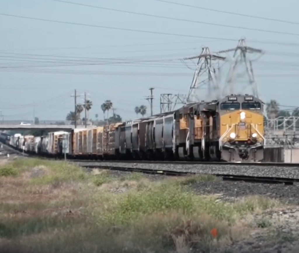 A train with a yellow and gray engine and cars passes by on tracks next to a grassy area, with power lines and palm trees in the background.