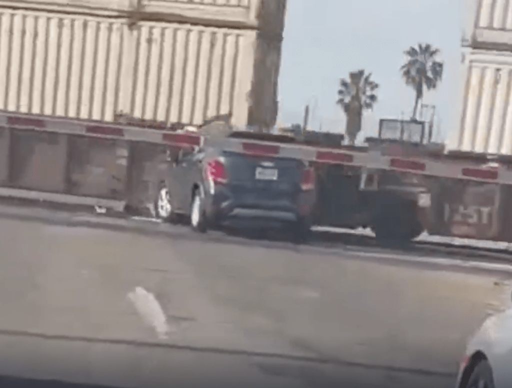 A dark-colored SUV is stuck on railroad tracks as a train approaches, its rear bumper hitting the side of a cargo car.