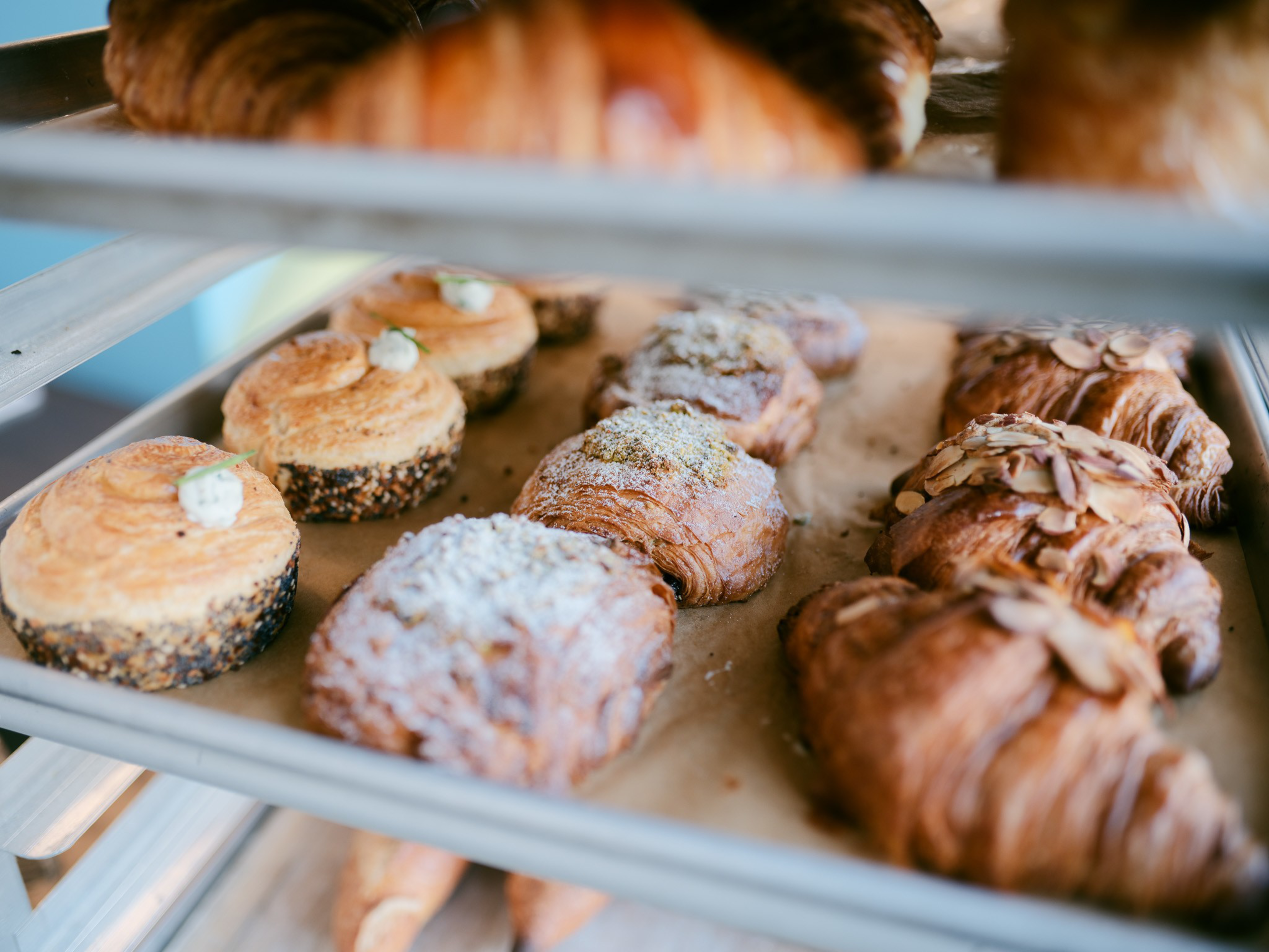 A tray of assorted baked pastries including croissants topped with sliced almonds, powdered sugar-dusted pastries, and round seeded buns with small dollops of cream.