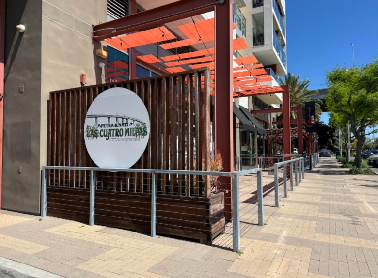 A small business, with a round sign on a pallet that forms a wall over a planter. The sign reads: Las Cuatro Milpas over the background of a bridge. 