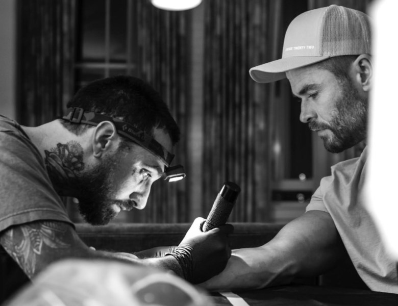 One man bends low over another man's arm. Tattoos are visible on his arm and neck as he works on a tattoo for the client, actor Chris Hemsworth.