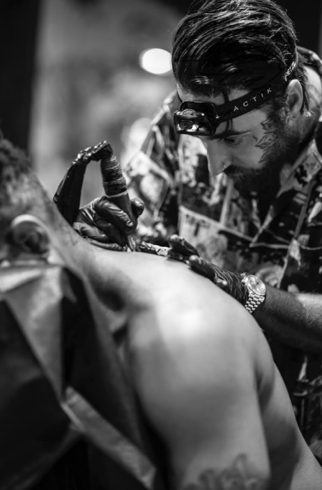A man with a tool in his hand inks a client's back. The client is lying face down on a chair, shirtless, and a tattoo can be seen on his left arm.