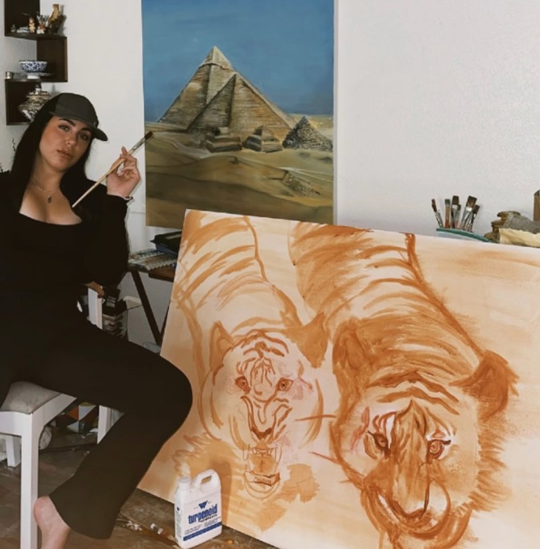 A female artist dressed casually in a cap leans back in a chair beside a large canvas showing two painted tigers. Another painting on the wall shows pyramids rising above desert terrain against a blue sky.