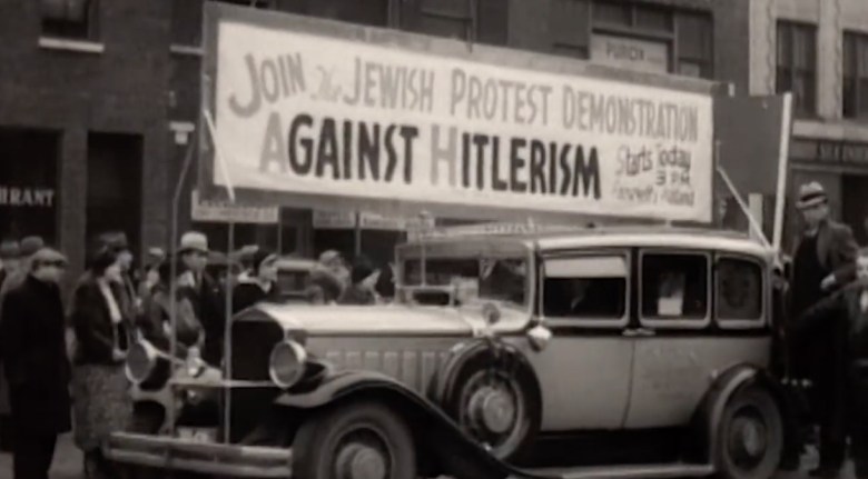 Archival footage of a U.S. protest against Hitler with a banner over a car proclaiming "Join the Jewish Protest Demonstration Against Hitlerism, starts today"