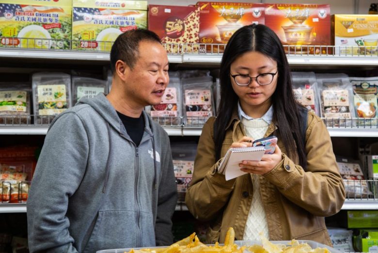 Two people stand in a store aisle; one person speaks while the other writes notes in a notepad. Various packaged goods are displayed on shelves behind them.