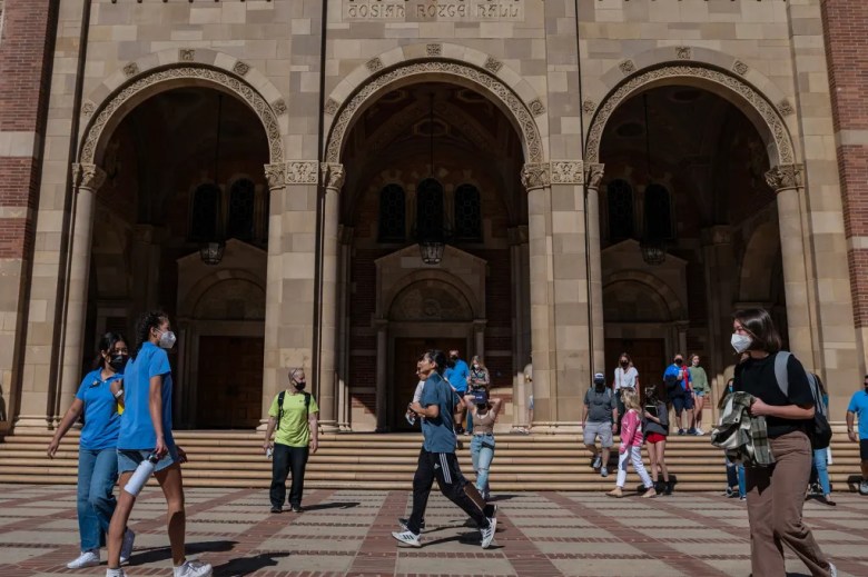 People walk pass a large red and brown brick building with three arch entrances.