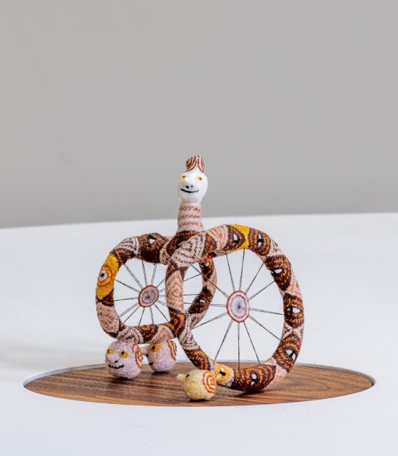 A small whimsical sculpture featuring two large wheel-like shapes and a smiling, snake-like figure with a white face, displayed on a round wooden base against a plain background.