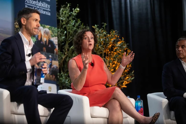 A panel discussion on a stage shows three speakers seated in white chairs. At center, a person in a bright orange dress speaks animatedly, hands raised mid-gesture, while wearing a lapel microphone. To the left, another panelist in a dark suit holds a water bottle, listening, and to the right, a third panelist looks on. A backdrop with greenery and a vertical banner about affordability and rural California is visible behind them.