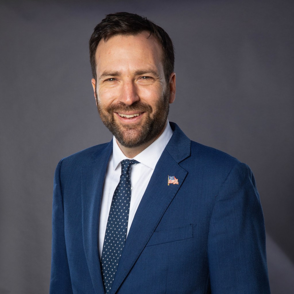 A portrait of a person with short brunette hair and wearing a blue suit with a white shirt.