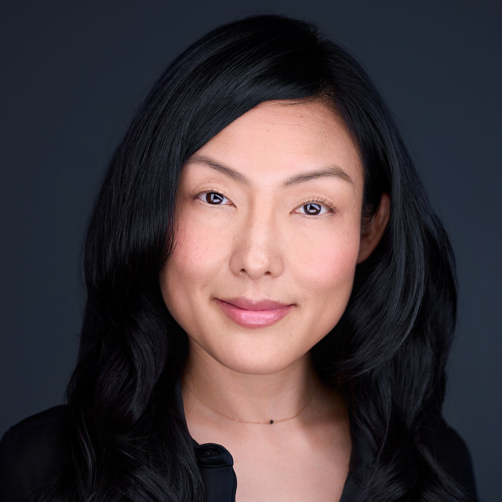 A close-up portrait shows a person with long, dark hair looking directly at the camera with a neutral, composed expression. The background is softly blurred, and the lighting is even and flattering, highlighting facial features without harsh shadows.