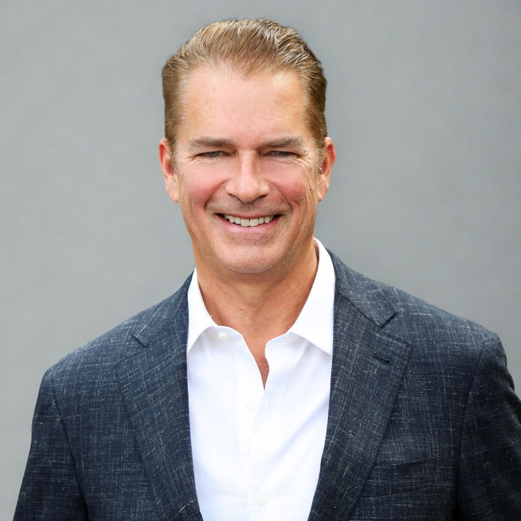 A portrait of a person with short blonde hair and wearing a dark gray suit with a white shirt.