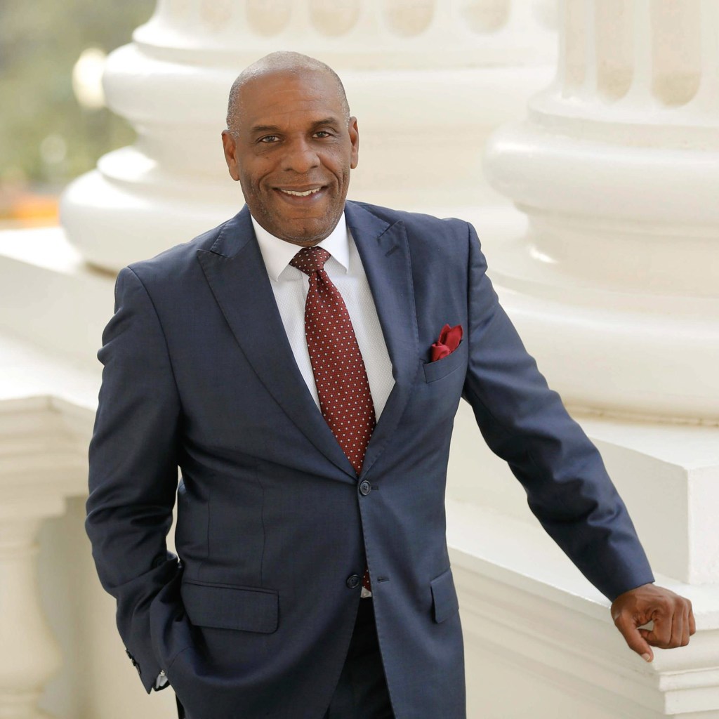 A portrait of a person wearing a blue suit with a red tie and they rest their right hand on a white pillar.