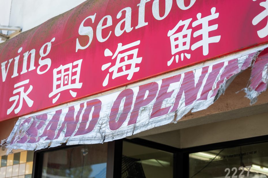 A torn "Grand Opening" banner hangs below a red storefront sign with English and Chinese text at a seafood store.