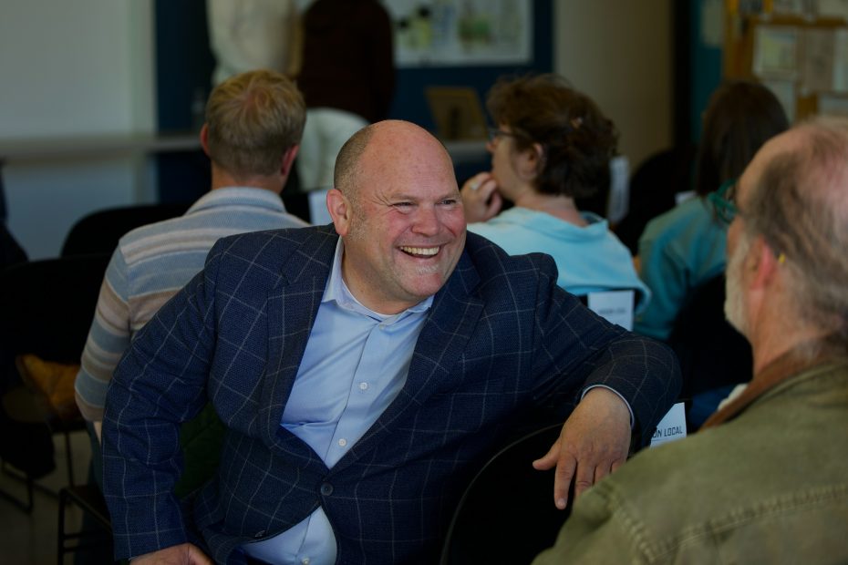 A man in a blue checked suit jacket smiles and talks with another person while seated among a group of people in a meeting room.