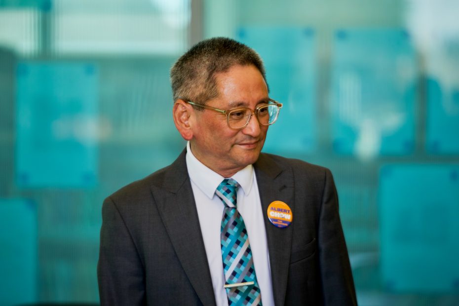 A man in a suit and glasses stands indoors, wearing a turquoise patterned tie and a button that reads "Alberta Checks.