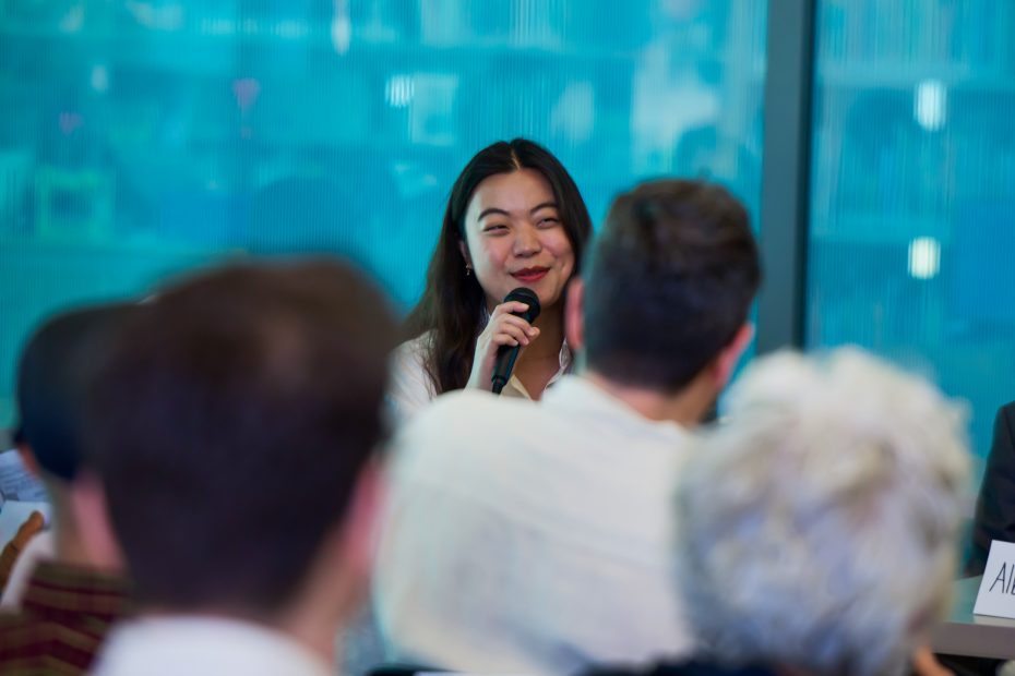 A woman holds a microphone and speaks at a panel discussion, facing an audience with several people visible from behind.