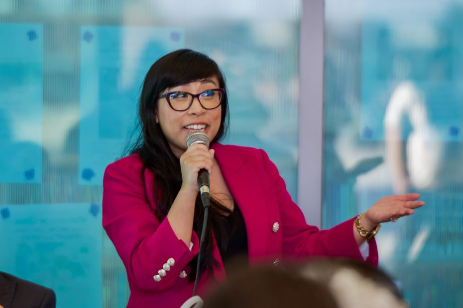 A woman in a bright pink blazer speaks into a microphone, gesturing with her hand during an indoor event.