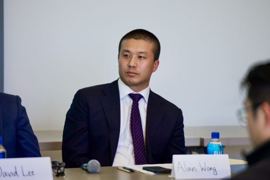 A man in a suit sits at a table during a panel discussion, with a microphone, water bottle, and name cards labeled "Alan Wong" and "David Lee" visible.