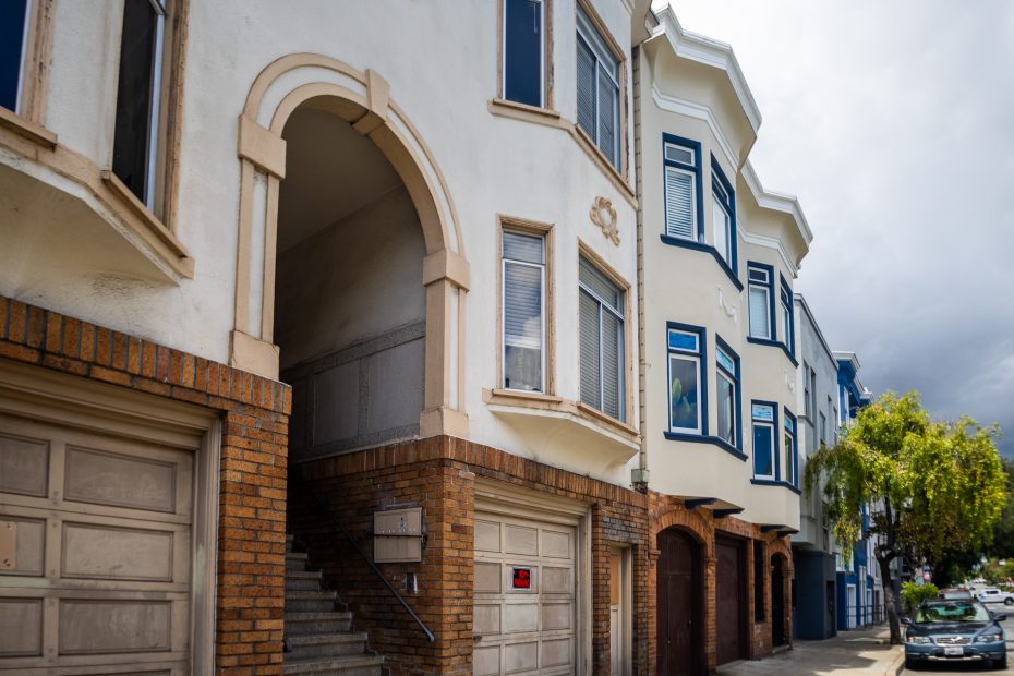 A row of multi-story residential buildings with garages at street level and a staircase leading to an arched entryway. Trees and parked cars are along the sidewalk.