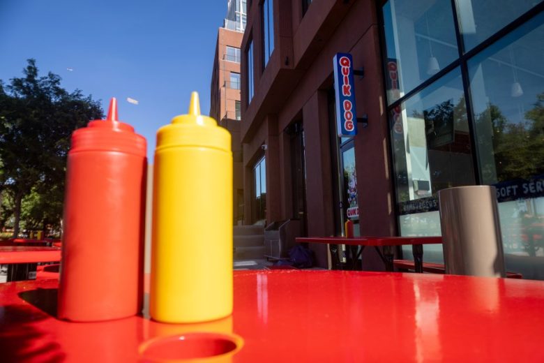 Red and yellow condiment bottles sit on a red outdoor table near a building with a "QUK DOG" sign in the background.
