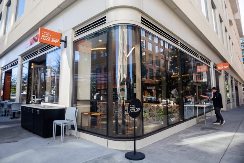 Street view of a modern pizza shop on a corner with large windows, outdoor seating, and a person entering at the door.