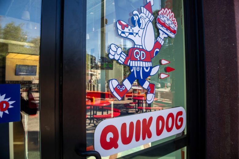 Glass door with a colorful cartoon dog holding a burger and wearing a "QD" shirt; text below reads "QUIK DOG." Red chairs and tables visible inside.