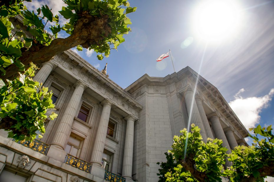 A stone government building with columns and a flag flying, seen from below on a sunny day, with green trees framing the view.