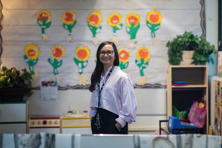A person wearing a white button-up shirt and black-framed eyeglasses stands in the center of a classroom, with floral decor in the background, and two rows of brightly colored paper sunflowers pinned to a white wall.
