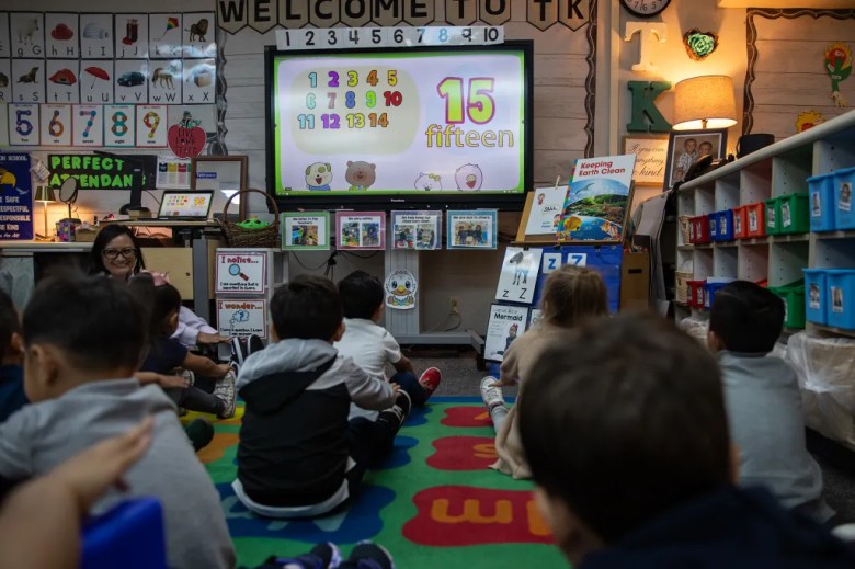 An over-the-shoulder view of a young student sitting cross-legged on the floor on a carpeted area in a brightly colored, decorated classroom, with a large television screen with a math problem displayed on it.