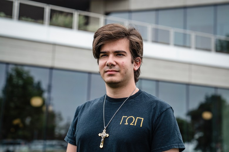 A young person stands outdoors in front of a modern building with large glass windows and metal railings. They wear a dark T-shirt with Greek letters on the chest and a necklace with a cross pendant. Their hair is medium-length and swept to the side, and they look slightly off-camera with a neutral expression.