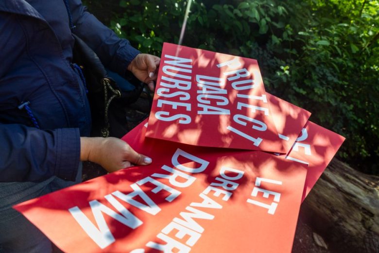 A person holds several red protest signs with white text reading messages supporting DACA, nurses, and Dreamers, outdoors with greenery in the background.