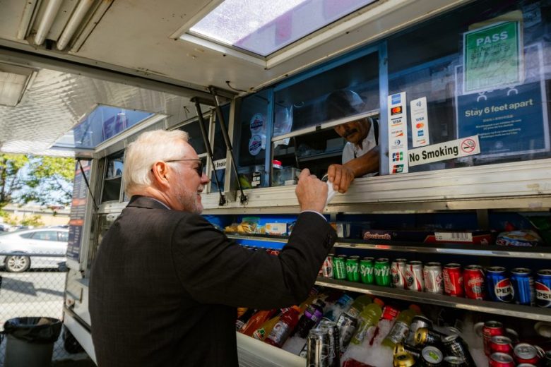 A man in a suit buys food from a vendor at a food truck window stocked with various bottled and canned drinks.