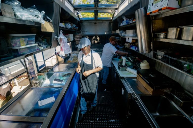 Two people work inside a food truck kitchen, preparing food and organizing supplies, with metal counters and shelves lining the space.