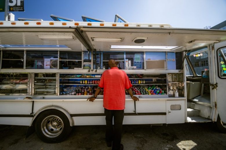 A man in a red shirt stands at the window of a white food truck lined with drinks and snacks on a sunny day.