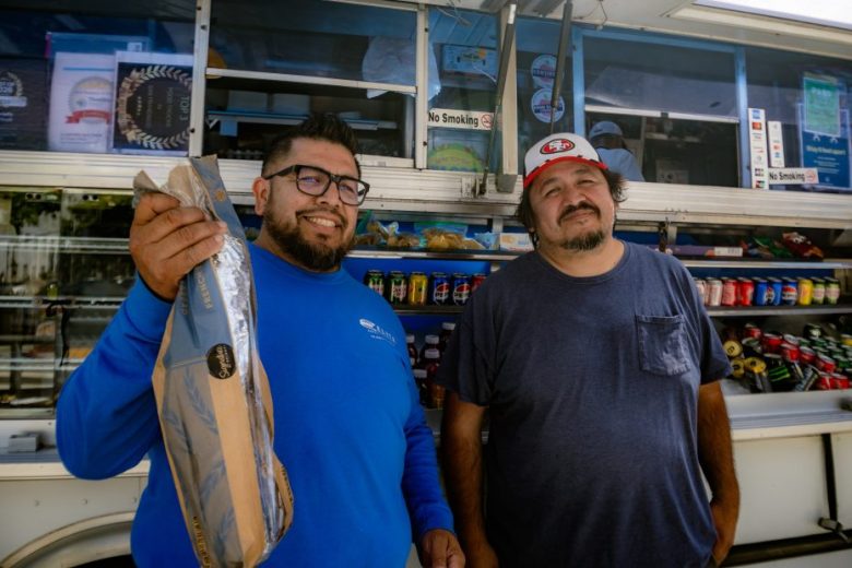Two men stand in front of a food truck; one holds a baguette and smiles, while the other stands with hands in his pockets. Various drinks and snacks are visible inside the truck.