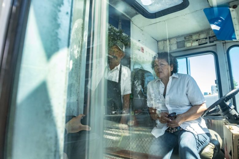 Two people are inside a vehicle; one is seated at the wheel holding a phone, while the other stands nearby. The scene is viewed through a reflective glass window.