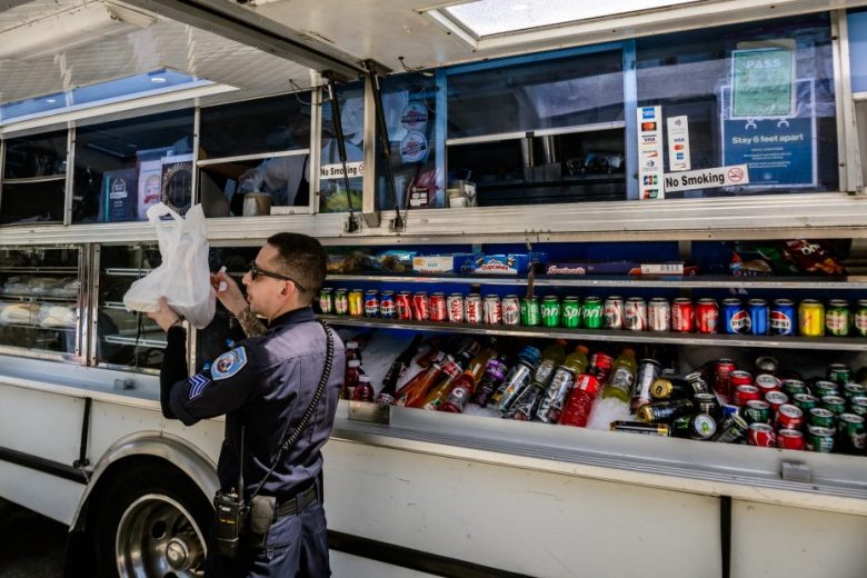 A uniformed officer receives a plastic bag of food from a food truck, with an array of sodas and drinks displayed in the window.