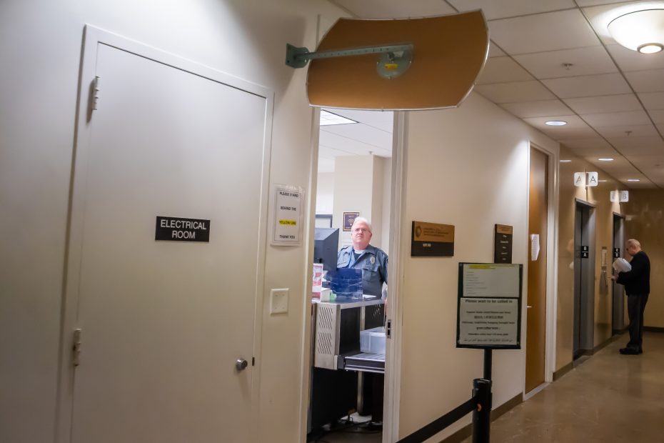 A security guard sits at a reception desk in a hallway near an “Electrical Room” door, with a broken sign hanging from the ceiling and another person using a phone in the background.
