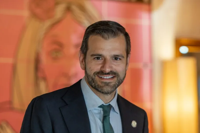 A man in a suit and tie stands indoors, smiling at the camera, with a pink mural featuring a woman's face in the blurred background.