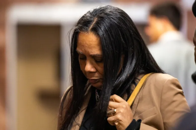A woman with long dark hair looks down while standing indoors, wearing a brown blazer and carrying a tan bag.