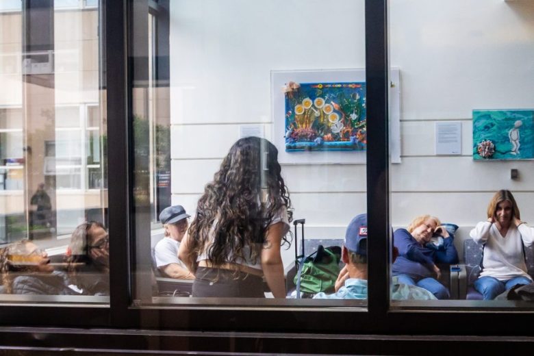 A group of people sit and wait in a room with artwork on the walls, viewed through a glass window. One woman stands while others are seated, some appearing to rest or talk.