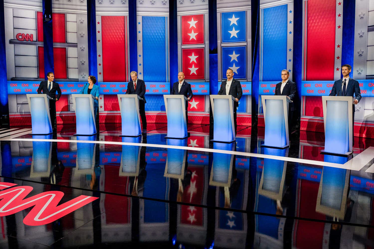 Seven candidates stand at blue-lit podiums on a CNN stage with a red and blue backdrop.