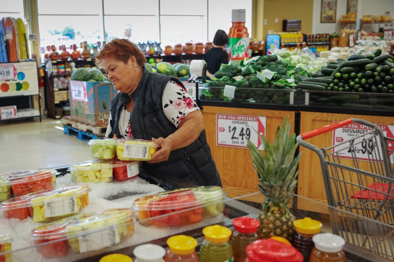 An older individual stands in a grocery store near a display of pre-packaged fruit containers on ice, selecting a pack of watermelon and other mixed fruits. The person is wearing a floral shirt under a dark vest. Behind them are avocados on sale, with a price sign for $2.49 per avocado. A shopping cart with a pineapple inside is visible nearby, along with other fresh produce such as cucumbers.