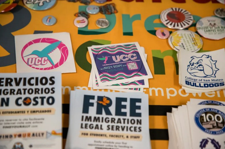 A close-up view of a table covered with colorful informational materials and stickers related to immigration services and campus organizations. In the center are square stickers for the Undocumented Community Center (UCC), featuring a stylized bird and wavy background. To the left is a pink and teal UCC hummingbird sticker, and to the right are College of San Mateo Bulldogs stickers. Various round buttons, brochures about free immigration legal services, and Spanish-language flyers are spread across the table.