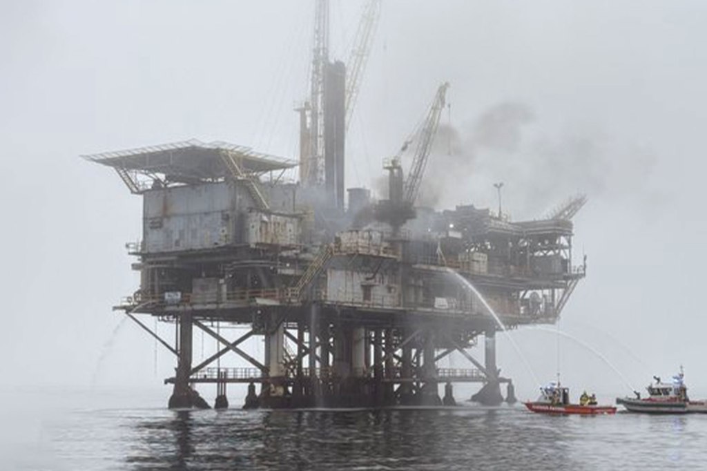 Oil platform on fire with two boats spraying water onto it.