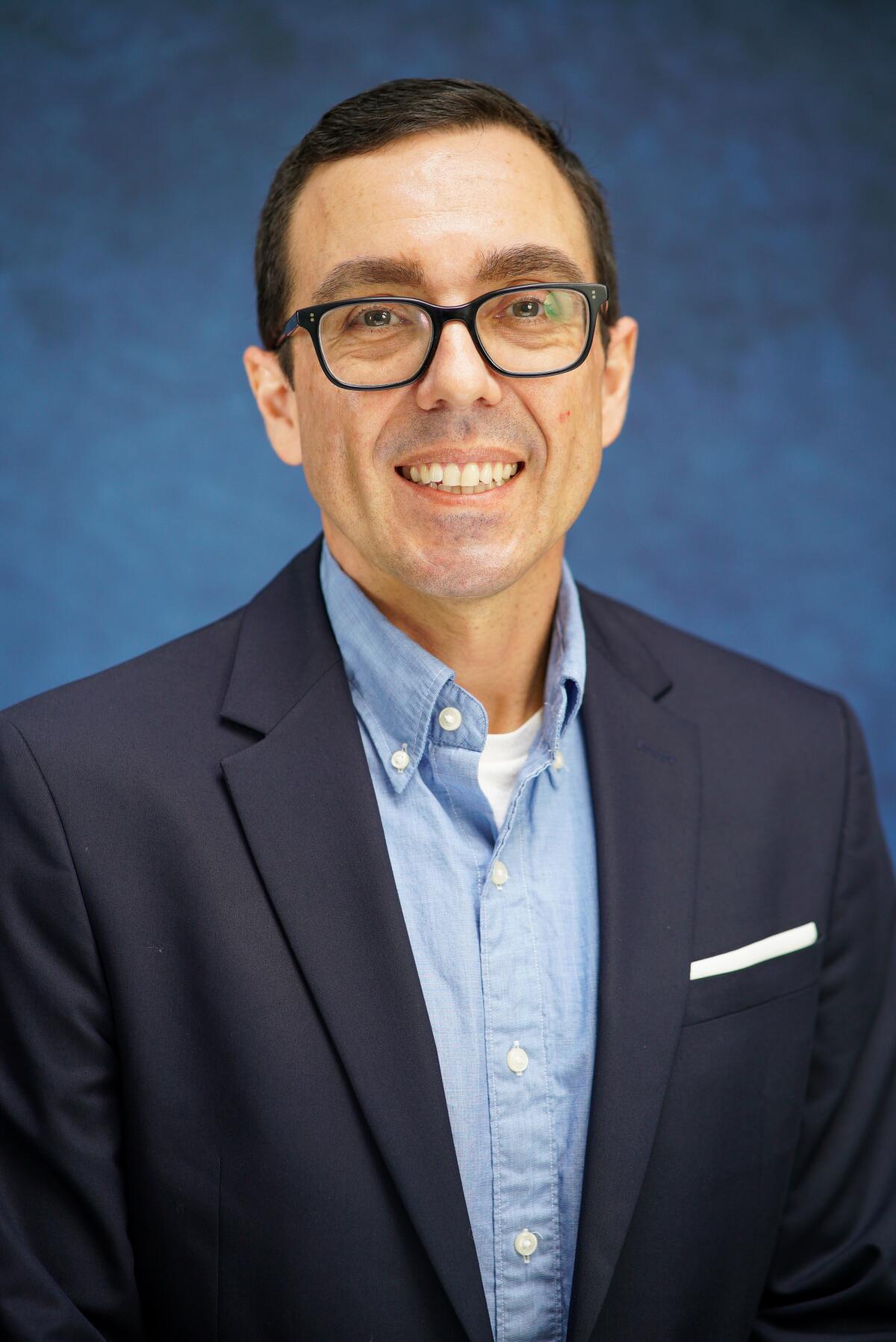 Morgan Oyler, a City Council candidate, in front of a blue background wearing black suit jacket, light blue button-up shirt. 